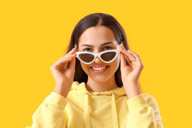 Güneş gözlüklü genç kadın ve sarı arka planda kapüşonlu, yakın plan.