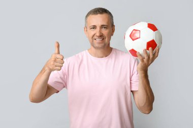 Futbol topuyla yakışıklı, olgun bir adam gri arka planda baş parmak hareketi yapıyor.