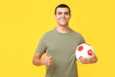 Futbol topuyla yakışıklı genç adam sarı arka planda baş parmak hareketi gösteriyor.