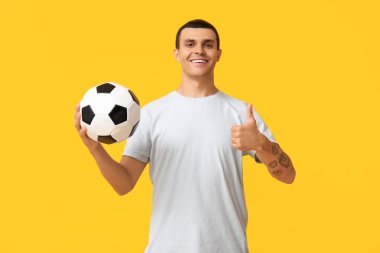 Futbol topuyla yakışıklı genç adam sarı arka planda baş parmak hareketi gösteriyor.