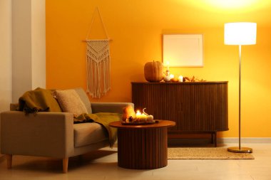 İçinde gri kanepe, yanan mumlar ve ahşap mobilyaların üzerinde balkabakları olan karanlık oturma odasının içi.