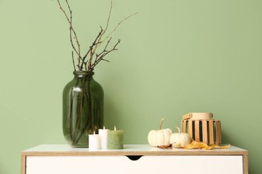 Yeşil duvarın yanında beyaz çekmeceler, balkabakları, mumlar ve ağaç dalları