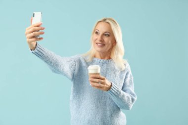 Elinde kahve olan olgun bir kadın mavi arka planda selfie çekiyor.