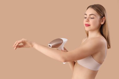 Güzel genç bir kadın, kahverengi arka planda modern fotoepilatörle kollarına epilasyon yapıyor.