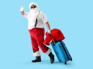 Noel Baba bir çanta dolusu hediyeyle ve mavi arka planda el sallayan bavulla