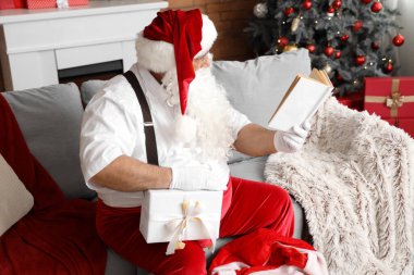 Noel Baba çanta ve Noel hediyesi kutusuyla oturma odasında kitap okuyor.