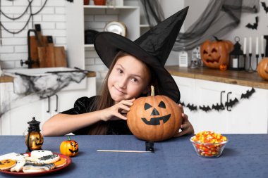 Cadılar Bayramı 'nda cadı gibi giyinmiş tatlı küçük kız mutfaktaki masada kurabiye ve şekerle.