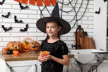 Cadılar Bayramı süslemeleri için mutfakta balkabağı ve şekerle cadı gibi giyinmiş şirin küçük kız.