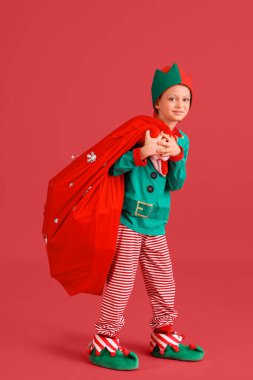 Kırmızı arka planda Noel Baba çantası olan elf kostümlü şirin bir çocuk.