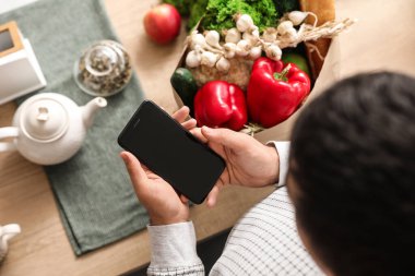 Bakkal çantalı adam mutfaktaki masada cep telefonu kullanıyor.