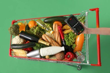 Yiyecek dolu alışveriş arabası ve yeşil arka planda ödeme terminali olan kadın eli.