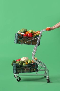 Yeşil arka planda içi taze sebze dolu alışveriş arabasını iten kadın.