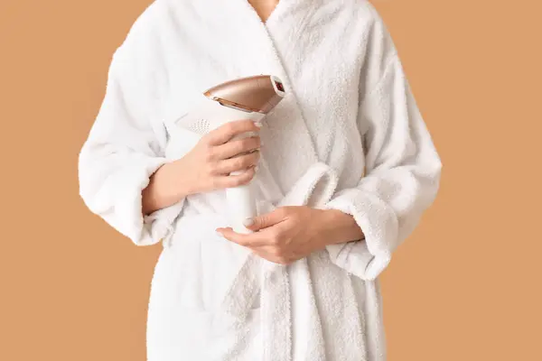 Beautiful young woman in bathrobe with photoepilator on brown background
