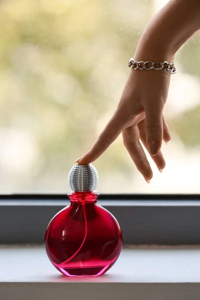 Female hand with bottle of elegant perfume on windowsill at home