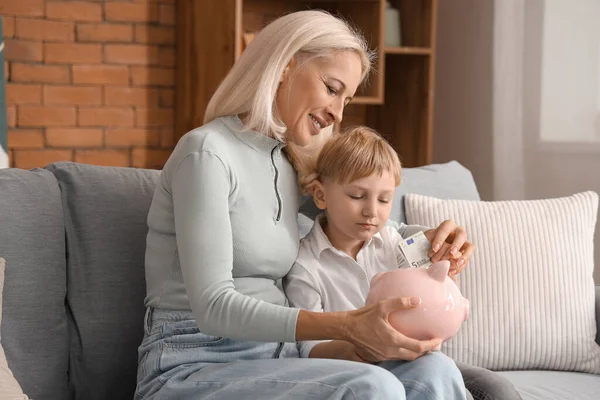 Little boy with his grandmother putting money in piggy bank at home