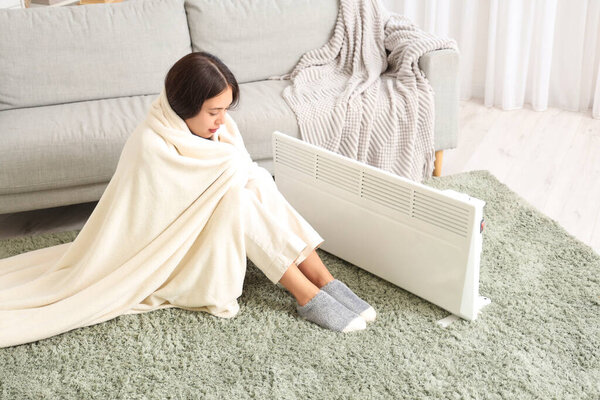 Pretty young woman in blanket warming near electric heater in living room