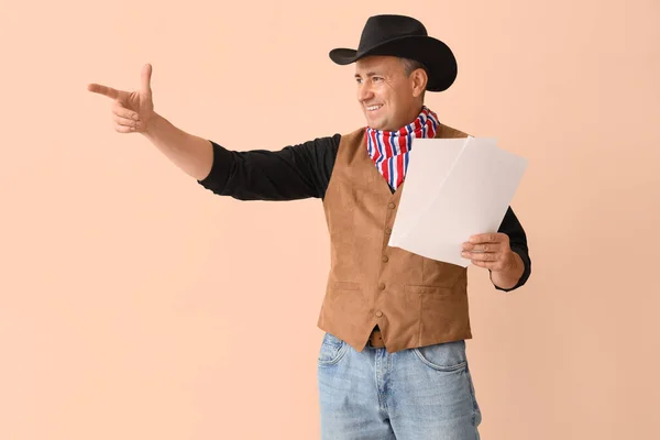 Mature actor dressed as cowboy with film script pointing at something on beige background