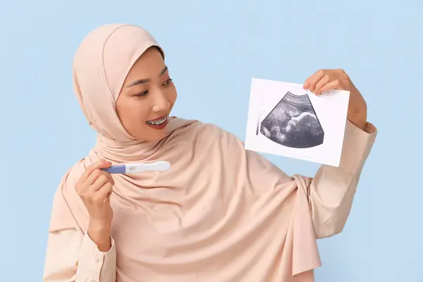 Young Muslim woman with pregnancy test and sonogram image on blue background