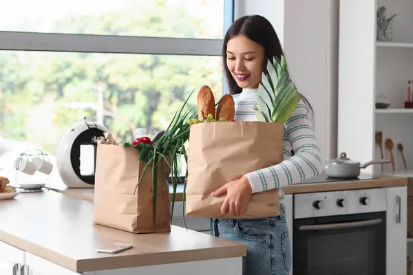 Young Asian woman with mobile phone and shopping bags full of fresh food at table in kitchen