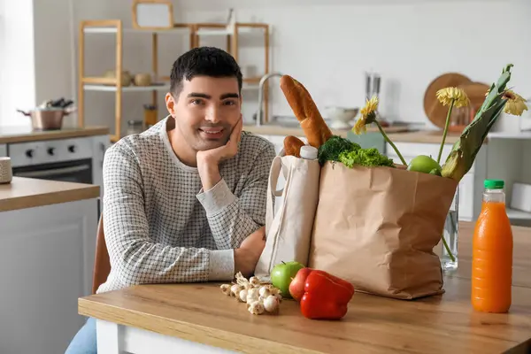 Young man with grocery bags at table in kitchen