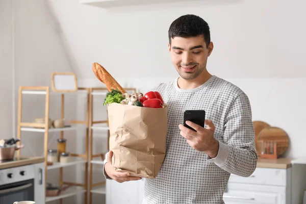 Young man with grocery bag and mobile phone in kitchen