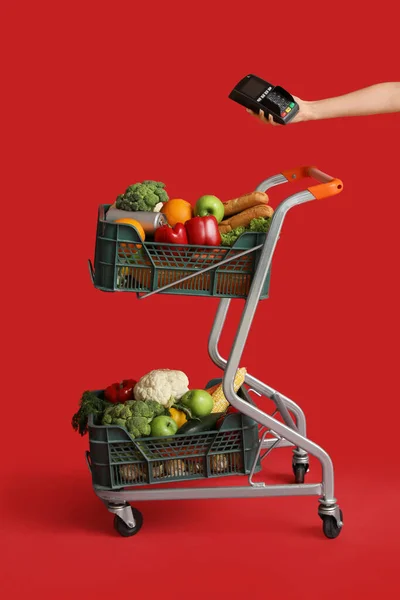 Shopping cart full of food and female hand with payment terminal on red background
