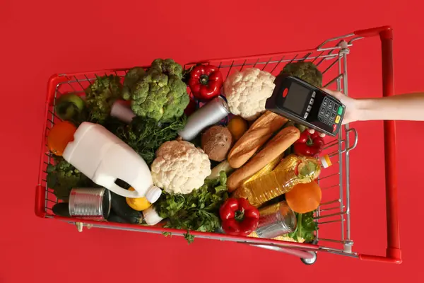 Shopping cart full of food and female hand with payment terminal on red background