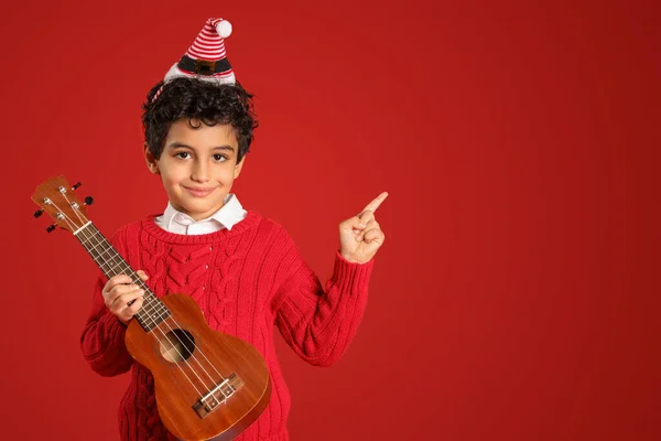 Cute Little Boy Santa Hat Guitar Pointing Something Red Background — Stock Photo, Image