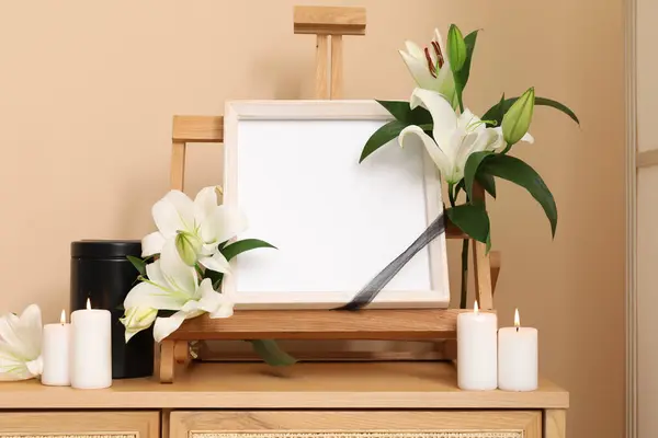 Blank funeral frame with lily flowers, mortuary urn and burning candles on wooden cabinet against color wall