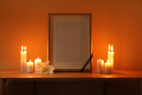 Blank funeral frame, burning candles and lily flower on wooden table in dark room
