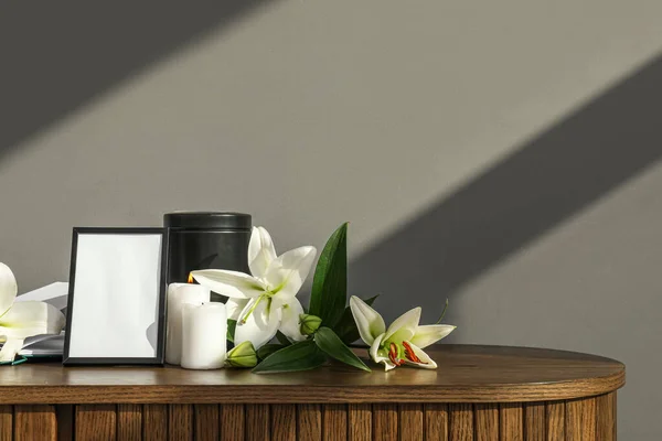 Blank funeral frame, mortuary urn, burning candles and lily flowers on wooden table near grey wall