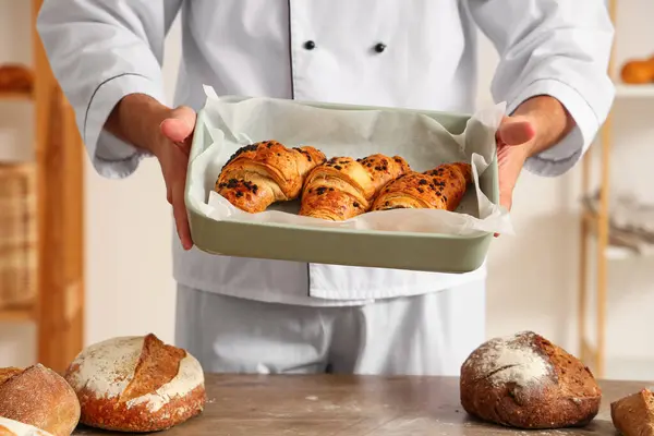 Handsome young chef holding baking dish with fresh croissants in bakery
