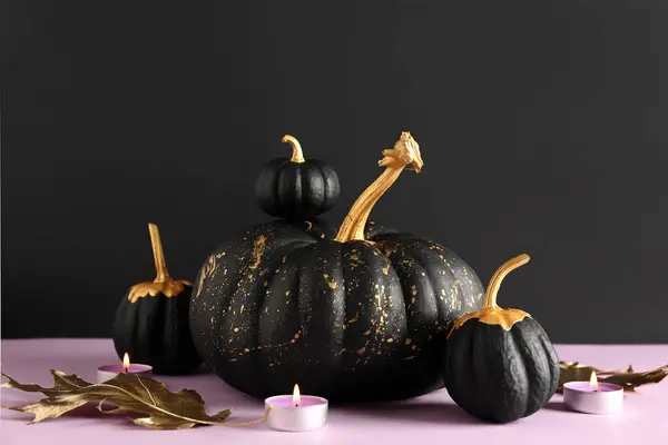 Painted pumpkins, candles and golden leaves on purple table near black wall