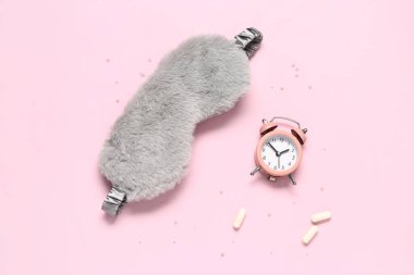 Şık bir uyku maskesi, çalar saat ve pembe arka planda haplar.