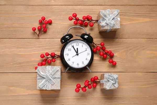 Christmas composition with alarm clock, red berries and gift boxes on brown wooden background