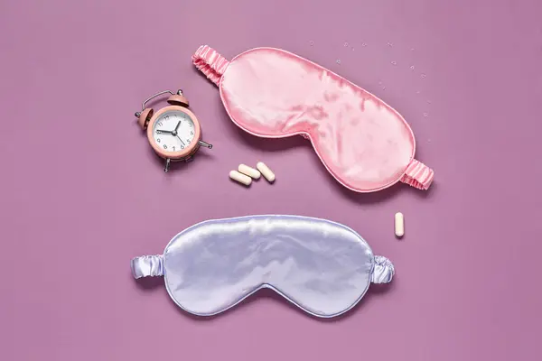Stylish sleep masks, pills and alarm clock on color background