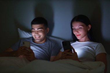 Geceleri yatak odasında cep telefonu kullanan genç bir çift.