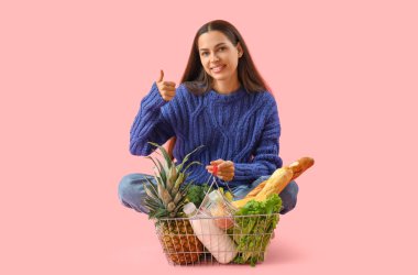 Alışveriş sepeti olan genç bir kadın pembe arka planda baş parmağını kaldırıyor.