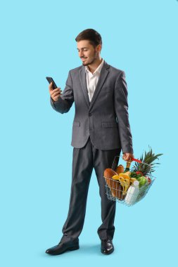 Mavi arka planda alışveriş sepeti ve cep telefonu olan genç bir adam.