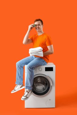 Genç adam turuncu arka planda çamaşır makinesinde oturuyor.