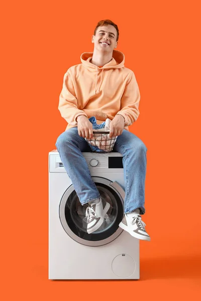 Young man with laundry sitting on washing machine against orange background