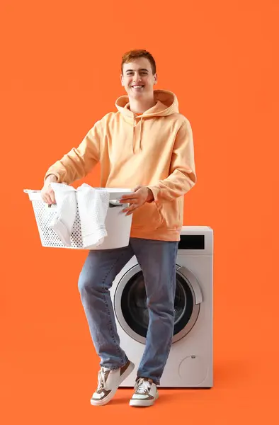 Young man with laundry near washing machine on orange background