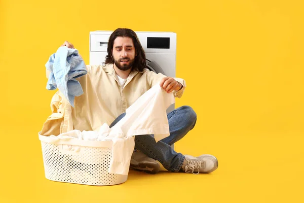Young man with laundry basket near washing machine on yellow background