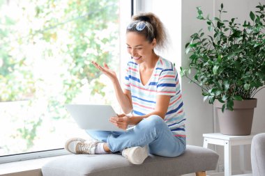 Evde pencerenin yanında oturmuş bilgisayar başında sohbet eden olgun bir kadın.