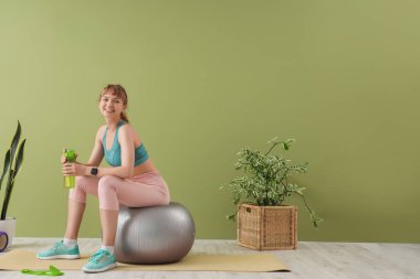 Elinde bir şişe su olan genç sporcu kadın yeşil duvarın yanında fitness topunun üzerinde oturuyor.