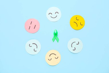 Farklı duygulara sahip kağıt yüzler ve mavi arka planda yeşil farkındalık kurdelesi. Ruh Sağlığı Günü