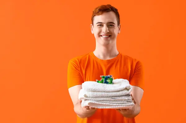 Young man with laundry capsule and towels on orange background