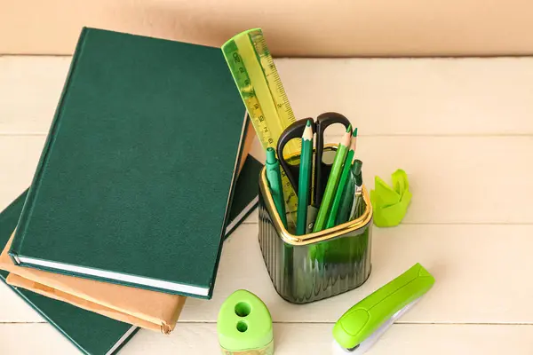 Holder with different stationery and books on light wooden desk, closeup
