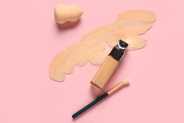 Bottle of makeup foundation with sample, brush and sponge on pink background
