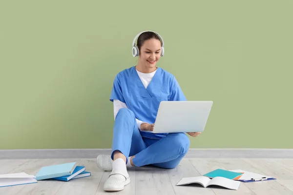 Female medical intern in headphones studying with laptop near green wall